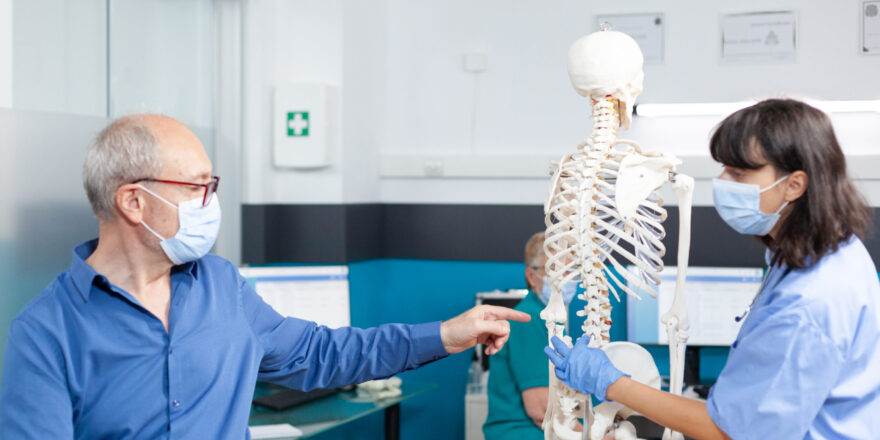 Patient and nurse with face masks looking at spine bones on human skeleton for osteopathy examination and physical recovery. Assistant showing spinal cord to old man during pandemic