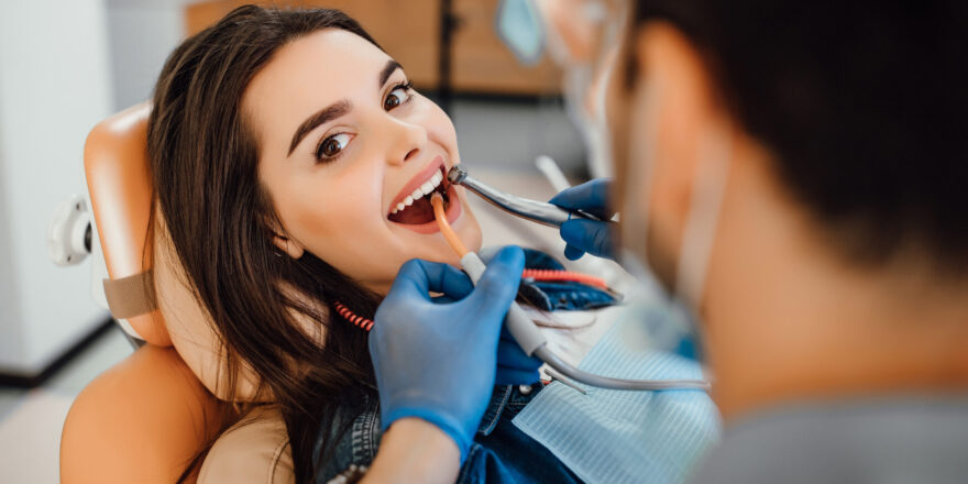 Young female patient visiting dentist office. Beautiful woman with healthy straight white teeth sitting at dental chair with open mouth during oral checkup while doctor working at teeth.